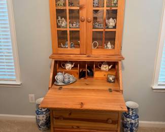Broyhill Pine Secretary Desk with Hutch (Bureau Bookcase) A charming country-style two-piece pine secretary desk with hutch, featuring: Top Hutch: Double glass-panel doors with decorative wood mullions, displaying collectible teapots and figurines on interior shelves. Crowned with an ornate broken-pediment bonnet top with carved floral/scroll detail. Secretary Section: A fold-down writing surface (slant-front drop lid) with interior cubbies/pigeonholes for organization, currently displaying small teapot collections. Base: Three drawers with round wood knob pulls, sitting on bun feet. Finish: Natural honey/amber pine with visible wood grain and knots.