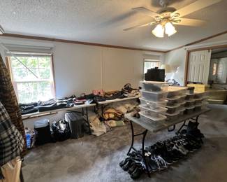 Wide view of the primary bedroom. There are piles of purses and other bags, and a table covered in shoe boxes. More shoes rest under the table. There are also piles of linens and comforters.