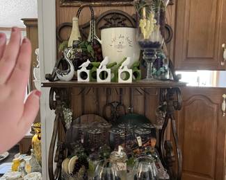 A wooden drink cabinet, with spaces for wine glasses to hang. Currently, this cabinet is covered in wine memorabilia, including hand-painted wine glasses, decanters, and items with a grape theme.