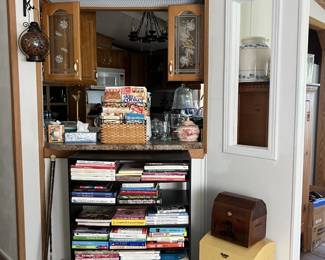In the foreground: a bookshelf filled with how-to manuals and cookbooks. Two small storage chests stack atop each other. In the background: a window into the kitchen. On top of a large shelf is a variety of vintage collectible tins, containers, and napkin holders.