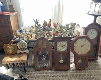 Part of a large collection of clocks. Two of the pictured are small and decorative, while the picture also includes four grandfather clocks. In the background, there is a table covered in small animal figures.