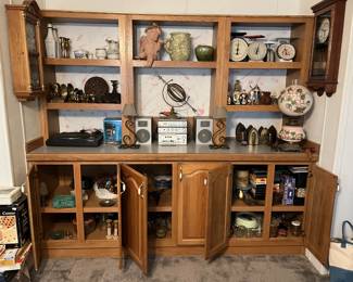 A large wooden display case filled with a variety of objects, including vintage scales, statues and figurines, a collection of brass irons, a pile of VHS tapes, and more.
