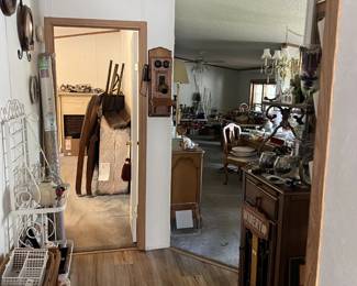 A view from the hallway of this home. It peeks into two rooms. The left is a bedroom, full of furniture and clothes. The right is a dining and living room. From this angle, you can see furniture, decorations, and some dishware.