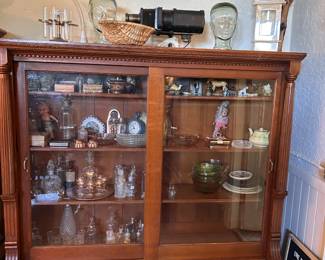 Cabinet display of bottles, clocks, small boxes, glassware, decanters, plates, figurines, green glass, and decorative curiosities.