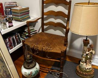 A vintage ladder back chair with damaged rush seat. Antique Royal Dux shepherd and boy lamp. Antique hand-painted oil lamp.  First edition children’s books.