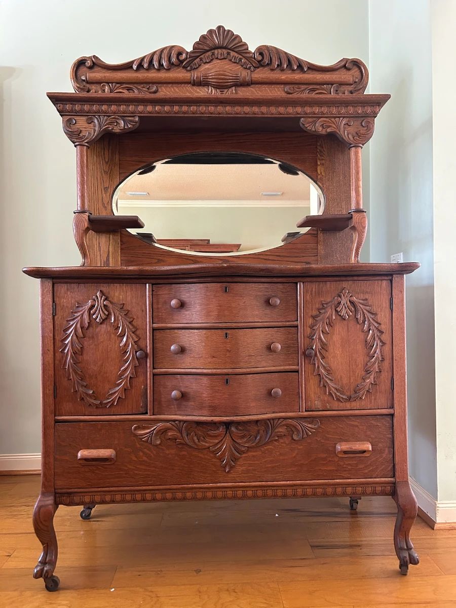 Beautiful antique solid oak hutch featuring intricate carved details, an elegant oval mirror, and rich wood grain throughout. Includes three center drawers, two side cabinets, and a large lower drawer for ample storage.