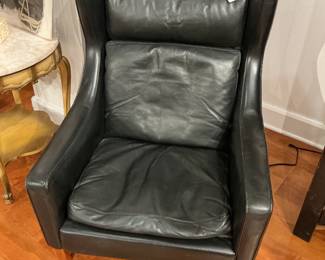 Danish Modern Style Black Leather Wingback Lounge Chair Description: A striking mid-century modern inspired high-back lounge chair upholstered in supple black top-grain leather. Borrowing design cues from iconic Scandinavian wingbacks, this piece features a gently contoured, enveloping shell silhouette with sleek sloped arms. It features a loose seat cushion and a matching plush lumbar/back cushion, topped with a distinctive, rectangular bolster headrest pillow held in place by integrated leather counterweight straps over the back. The frame is grounded by beautifully tapered, round solid wood dowel legs in a warm, natural honey-brown finish that provides a sharp, organic contrast to the dark leather. Estimated Dimensions: Width: 32" Depth: 34" Height: 40" Seat Height: 17"