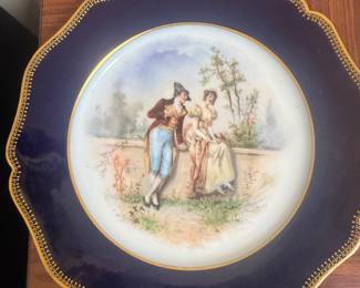 Limoge This porcelain cabinet plate features a hand-painted romantic scene with figures in 18th-century attire, set within a vibrant cobalt blue border decorated with intricate gold gilding