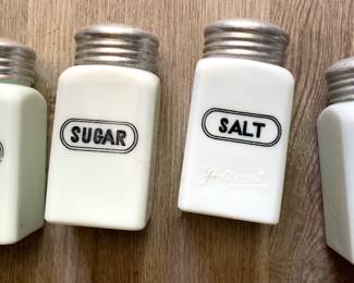 This set consists of vintage Hazel Atlas milk glass shakers featuring black shield graphics for pepper, sugar, salt, and flour.