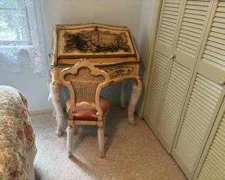 The most distinctive piece in the collection. A hand-painted Italian Venetian-style secretary desk (bureau à abattant) in cream and gilt with chinoiserie landscape scenes on the fall-front lid. Serpentine front drawer below, cabriole legs, decorative tassel key. The legs are wrapped in bubble wrap suggesting it was recently moved or is being protected. Accompanied by a matching painted and gilt side chair with cane back panel and coral/salmon upholstered seat. This is a specialty antique — likely 1960s Italian export reproduction, but the hand-painting makes it genuinely decorative and collectible.