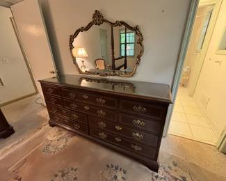 Large mahogany triple dresser with 9 drawers and brass bail pulls, bracket feet, and a glass top protector. Accompanied by a large ornate gilt-framed Rococo revival mirror with scrollwork and shell crest.
