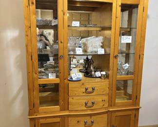 Large two-piece knotty pine china cabinet with three glass-front upper display doors, interior lighting, glass shelves with plate grooves, and mirrored back panels. Lower section has two drawers and two cabinet doors with a serpentine front. Antique brass bail pulls. Good condition with warm honey pine finish. Seats a full china service with room to spare.
