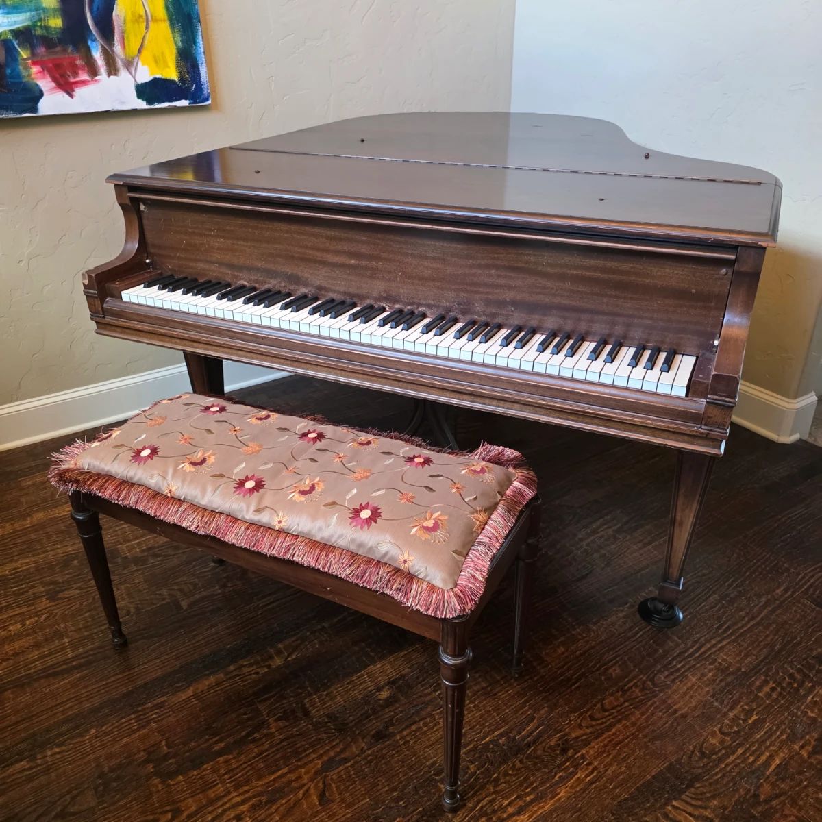 This vintage grand piano features a warm wood finish and is accompanied by a matching bench with a decorative floral cushion and fringed trim.