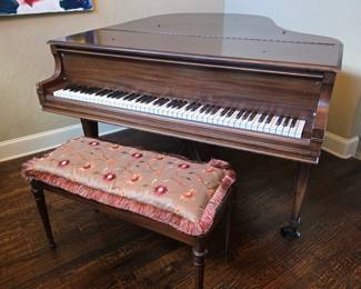 This vintage grand piano features a warm wood finish and is accompanied by a matching bench with a decorative floral cushion and fringed trim.
