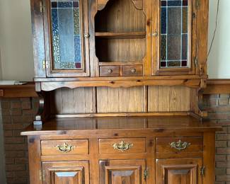 Early American Colonial style china cabinet / hutch. It features a two-piece construction consisting of a lower buffet storage base and an upper display deck accented by leaded, multi-colored stained glass door panels.
