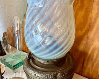 Featured is a stunning antique Victorian-era banquet/parlor lamp featuring an exquisite blue-white opalescent swirl glass shade with a beautifully ruffled rim. The lamp boasts a heavy, ornately cast metal base decorated with deep, intricate Victorian scrollwork and relief detailing. This piece has been tastefully converted to electricity, making it perfectly functional as a ready-to-use statement piece or accent light for a living room, entryway, or bedroom. A true collector's item in excellent vintage condition! Available firsthand at the estate sale