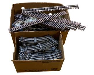 1153 - 2 Boxes O Scale Model Train Tracks (Description): Two boxes of O scale model train tracks. The collection includes both straight and curved track sections, providing a variety of pieces for building or expanding a model railroad layout. The tracks feature the characteristic three-rail design typical of O scale trains.