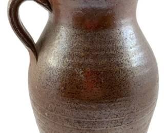 121 - Vtg Brown Stoneware Pitcher 10"x7"x6" (Description): Vintage brown stoneware pitcher featuring a classic bulbous form with a pinched spout and single loop handle. The pitcher displays a rich salt-glazed finish with natural color variations ranging from warm brown to rustic orange tones. Horizontal incised banding adorns the lower portion of the body. Measures 10 inches tall by 7 inches wide by 6 inches deep.