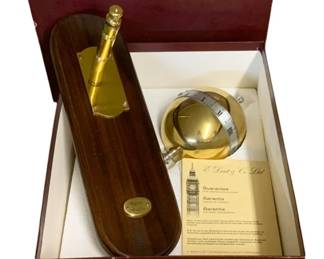 1134 - Dent London Clock in Original Box 16"x7" (Description): Dent London clock presented in its original burgundy presentation box with guarantee certificate. The set includes a gold-toned globe-style desk clock with Roman numeral markings on a silver-toned band, accompanied by a wooden desk stand with brassfittings and an engraved plaque. Made by E. Dent & Co. Ltd., the renowned London clockmakers known for creating Big Ben. Measures 16 inches by 7 inches.