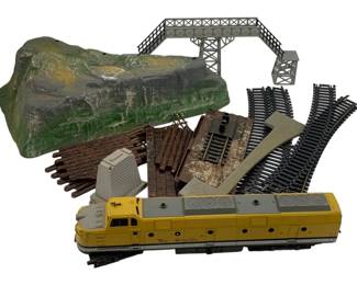 1004 - Lot HO Scale Train Items (Description): A lot of HO scale train items including a Rio Grande diesel locomotive in yellow and gray livery, assorted track sections, a signal bridge, terrain piece with rock formation, and various layout accessories including fencing, lumber loads, and scenic details.