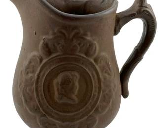 107 - Bennington Pottery Milk Pitcher (Description): Vintage Bennington pottery milk pitcher in brown stoneware featuring a molded medallion portrait design with decorative scrollwork and acanthus leaf motifs. The pitcher has a classic curved spout and ornate handle. Shows slight damage. Measures 8 inches by 8 inches by 4 inches.