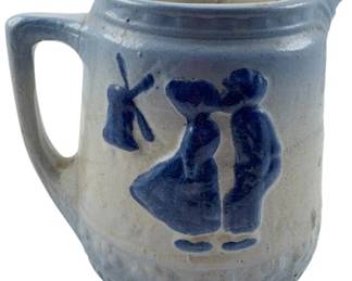 97 - Brush McCoy Salt Glazed Stoneware Pitcher (Description): Vintage Brush McCoy salt glazed stoneware pitcher featuring a blue and white color scheme with a Dutch-inspired design. The pitcher displays raised relief decoration depicting a kissing Dutch couple and windmill on the front. Measures 7 inches by 7inches by 4.5 inches.