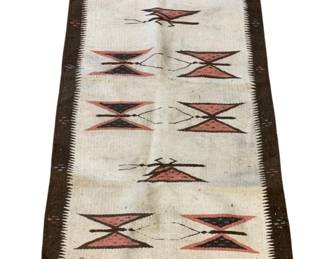 1144 - Vintage Native American Rug 60"x30" (Description): Vintage Native American rug featuring a cream or natural white ground with a dark brown border. The rug displays a traditional geometric design with repeating bow-tie or hourglass motifs in pink/coral, black, and brown tones arranged in vertical columns. The border features a distinctive comb or tooth pattern along the inner edge. Measures 60 inches by 30 inches.