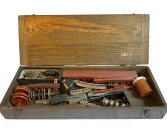 1062 - Gilbert Erector Set #7 in Wood Box 21"x2"x2" (Description): Gilbert Erector Set #7 housed in its original wooden box marked "No. 7" on the interior lid. The set includes various metal construction pieces, gears, and components typical of the classic Erector building system. Box measures 21" x 2" x 2".