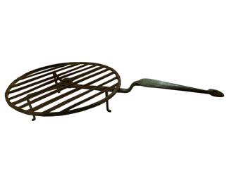 66 - Antique Hand Forged Wrought Iron Trivet (Description): Antique hand forged wrought iron rotating trivet featuring a round grated cooking surface with parallel bars, supported by three short legs. The trivet has a long handle with a hanging loop at the end. The piece displays a dark patina consistent with age and use. Measures 3 1/2" x 20" x 11".