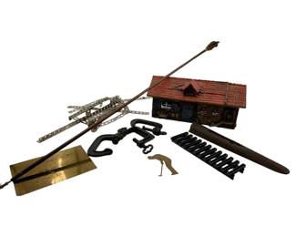 26 - Group Lot of Misc Decorative & Metal Items (Description): A group lot of miscellaneous decorative and metal items including a painted miniature log cabin model with red shingled roof, a decorative metal arrow, a metal erector set-style bridge or ladder structure, cast iron decorative elements including what appears to be a small fence section and C-clamp style pieces, a brass plaque with engraved text reading 'The Difference Between Men and Boys Is The Price of Their Toys,' a