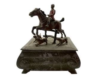 3 - Vtg Metal Box w/ Hunting Scene on Top (Description): Vintage metal box featuring an elaborate hunting scene on the lid. The figural top depicts a rider on horseback accompanied by two hunting dogs in mid-stride. The rider wears a red hunting jacket. The box itself rests on four small feet and features ornate decorative molding with scrollwork, egg-and-dart patterns, and acanthus leaf designs. The piece has a weathered patina throughout. Measures 6" x 5.25" x 4.25".