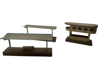 327 - Marklin Train Station Buildings 4"x3"x7" (Description): Marklin train station buildings featuring two vintage painted tin structures. The set includes a double-platform station with corrugated roof canopies supported by metal posts and a miniature bench detail, mounted on a rectangular base. The second piece is a control tower or signal box with observation windows and numbered panels on the front. Both pieces show characteristic vintage Marklin construction with painted surfaces