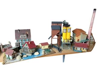 325 - Group Lot of HO Scale Train Buildings (Description): Group lot of train display items featuring an assortment of HO scale model buildings and structures. The collection includes a half-timbered European-style house, residential cottages with red roofs, a watermill with pond feature, industrial cement silos marked "Schwenk Zement," a large factory or mill building with corrugated siding, a brick smokestack, and various other architectural pieces arranged on a scenic