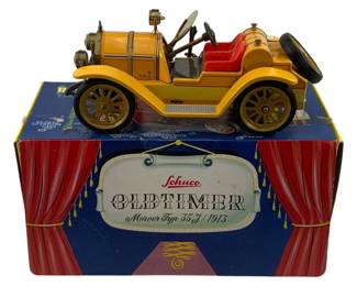 1182 - Schuco Oldtimer 1913 Mercer Typ 35J in Box (Description): Schuco Oldtimer 1913 Mercer Typ 35J model car in original box. This vintage tin toy features a yellow body with red interior seats, gold-tone spoke wheels, and detailed accessories including a spare tire mounted on the rear. The car is presented with its original decorative box featuring theatrical red curtain graphics and the Schuco Oldtimer branding.