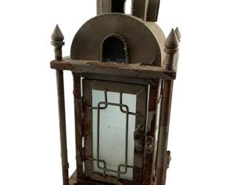 61 - Vintage Wrought Iron Lantern 16"x7"x7" (Description): Featuring an architectural design with decorative finials at the corners and an arched top with a carrying ring. The lantern has glass panels with ornamental ironwork detailing on the front door. The piece shows a weathered patina with areas of rust consistent with age and outdoor use. Rests on decorative claw feet. Measures 16 inches tall by 7 inches wide by 7 inches deep.
