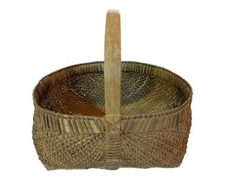 75 - Carolina Georgia 2 Tone Primitive Basket (Description): Carolina Georgia two-tone primitive basket with handle. This handwoven gathering basket features a traditional buttocks or melon shape with a sturdy bent wood handle secured through the center. The weave displays characteristic two-tone coloring with natural and darker brown tones throughout. The basket exhibits fine ribbed construction along the rim and tightly woven splint work on the body. Measures 13 inches by 15 inches