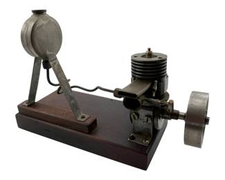 21 - Stirling Engine Model on Wood Base (Description): Stirling engine model mounted on a polished wood base. This display piece features a metal engine with finned cylinder head, flywheel, and elevated fuel reservoir connected by a support arm. The engine components have an aged patina consistent with vintage or handcrafted construction. Measures 6 1/2" x 9 1/2" x 4".