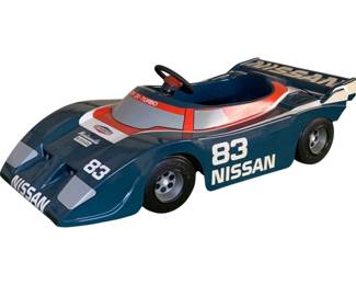 1089 - Nissan Electric Ride-On Race Car No Charger (Description): Nissan electric ride-on race car in blue, red, and silver racing livery featuring number 83 and Nissan ZX-Turbo graphics. This child's pedal-style car includes a steering wheel and rear spoiler. Sold without charger and noted as damaged. Measures 18" x 45" x 23".