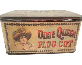113 - Vtg Dixie Queen Plug Cut Tobacco Tin (Description): Vintage Dixie Queen Plug Cut tobacco tin featuring a lithographed portrait of an elegant Victorian woman wearing a wide-brimmed hat. The tin displays decorative floral borders along the top edge and the brand name in bold red lettering. Shows considerable age-appropriate wear including surface rust, paint loss, and patina consistent with its age. Measures 3 inches by 6 inches by 3.5 inches.