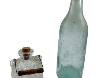116 - Vtg Group of 2 Glass Bottles 9.5" (Description): Vintage group of two antique glass bottles. The collection includes a clear pressed glass pineapple-shaped flask with a cork stopper and metal bail handle with wooden grip, and a pale aqua glass bottle with embossed text "This Bottle Not To Be Sold" featuring a porcelain and wire swing-top closure. The taller bottle measures 9.5 inches in height.