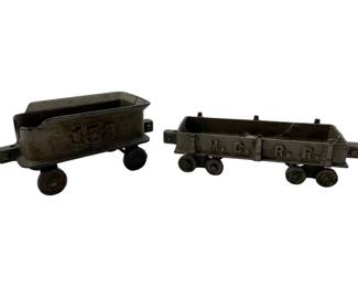 181 - 2 Vintage Cast Iron Gondola Train Cars (Description): Two vintage cast iron gondola train cars. One car is marked with the number 158, while the other features M.C.R.R. lettering on the side. Both cars feature open-top gondola bodies mounted on wheeled chassis with a dark oxidized patina consistent with age. Measurements: 2.5" x 6" x 2.5".