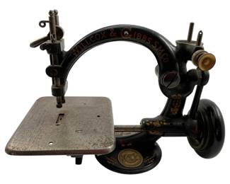 18 - Antique Willcox & Gibbs Sm Co Sewing Machine (Description): Antique Willcox & Gibbs Sm Co sewing machine featuring a cast iron body with black japanned finish and gold decorative scrollwork details. The machine displays the classic curved arm design characteristic of early Willcox & Gibbs models, with the company name prominently lettered across the top arch. Features include the original metal sewing plate with patent information, hand crank mechanism with wooden handle, and the