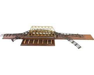 1099 - O Scale Train Bridge & Tracks Display Set (Description): O scale model train bridge and tracks display set. Features a brass-colored metal truss bridge mounted on a wooden base, accompanied by multiple sections of curved and straight track pieces mounted on walnut-toned wooden display boards. The set includes track switching components and appears to be a quality hobbyist display piece suitable for model railroad enthusiasts.