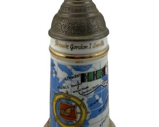 1083 - U.S. Military Beer Stein 10"x6" (Description): U.S. Military beer stein personalized for Private Gordon J. Smith. The porcelain stein features a decorative pewter lid topped with an eagle finial. The body displays a map of Europe including France, Germany, and Spain, along with military service ribbons and a Transportation Battalion insignia. Gold trim accents the rim and base. Measures 10" x 6".