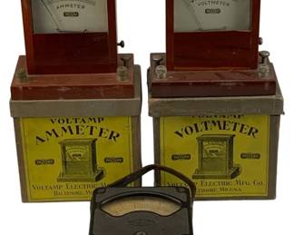 1091 - 3pc Lot Antique Volt Meters w/ 2 Boxes (Description): A collection of three antique volt meters, including two Voltamp Electric Mfg. Co. meters from Baltimore, MD housed in their original wooden cases with yellow lithographed boxes. The set includes a Voltamp Ammeter and a Voltamp Voltmeter, both featuring glass-fronted displays in mahogany-stained cases with metal binding posts. The third piece is a smaller portable meter in a black metal case with carrying
