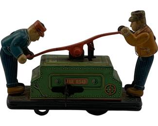 1120 - Tin Litho Wind-up Railroad Handcar Toy (Description): Vintage tin lithograph wind-up railroad handcar toy featuring two workers operating a pump-action handcar. The toy displays colorful lithographed details with a green handcar body marked "RAIL ROAD" on the front panel, and two figures in period railroad worker attire, one in blue and one in tan, pumping the lever mechanism. The piece sits on a black base with wheels for movement when wound. Measures 3 1/2" x 6" x 2".