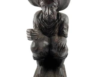95 - Vtg Handcarved Wooden Figurine Seated Man (Description): Vintage handcarved wooden figurine depicting a seated man wearing a wide-brimmed sombrero. The figure is carved in a folk art style, showing a bearded man in casual attire crouching on a rectangular base. The dark stained wood features visible chisel marks characteristic of hand carving. Measures 15.5" x 5.5" x 7".