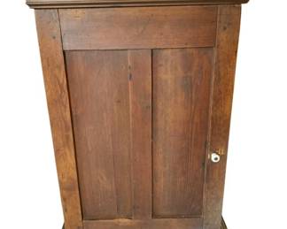 68 - 1 Door Wooden Hanging Primitive Cabinet (Description): A single door wooden hanging primitive cabinet featuring rustic pine construction with a warm, aged patina. The cabinet showcases a paneled door with a white porcelain knob, flanked by decorative molding at the crown and base. Brass hanging hardware is mounted at the top for wall display. The piece exhibits charming wear consistent with its primitive style. Measures 29" x 21.25" x 8".