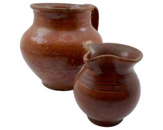 127 - Group of 2 Brown Clay Pitchers 5" (Description): Group of two brown clay pitchers featuring traditional rustic glazed earthenware construction. The larger pitcher has a simple rounded body with a single handle, while the smaller pitcher displays a ribbed decorative band around its midsection and a pinched spout. Both pieces exhibit a rich brown glaze characteristic of folk pottery traditions. Measures 5 inches.