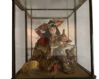 157 - Asian Doll in Glass Display 14"x12"x10" (Description): Asian doll displayed in a glass and wood display case. The intricately dressed figure wears elaborate traditional costume featuring rich brocade fabrics in red, gold, black, and silver tones with detailed embroidery and ornamentation. The doll is adorned with a traditional headdress and appears to be in a seated position surrounded by decorative accessories including a gold fan. The display case features a dark wood frame with glass